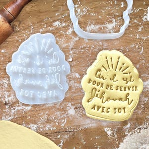 Könnte beinhalten: Ein weißer Plastik-Ausstecher mit dem Text "Que il est doux de servir Jehovah avec toi" in einem Sonnenstrahl-Design. Der Ausstecher befindet sich auf einer Holzoberfläche mit einem Nudelholz und einem Keks, der mit dem Ausstecher ausgestochen wurde.