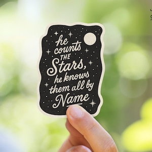 May include: A black sticker with white text that reads "he counts the Stars, he knows them all by Name." The sticker has a night sky design with stars and a moon. The sticker is held by a hand.