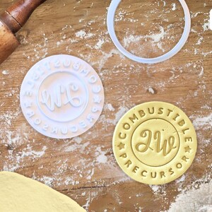 Op de afbeelding: Twee witte plastic koekjesvormen met het woord "Live" in een cursief schrift. De grotere vorm heeft de woorden "Combustible Precursor" rond de buitenkant van de cirkel. De kleinere vorm heeft de woorden "Combustible Precursor" rond de buitenkant van de cirkel.