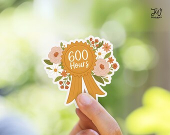 May include: A sticker featuring a gold-colored award ribbon with the text "600 Hours" in a white, elegant font. The ribbon is adorned with a floral arrangement of peach and white flowers, and green leaves. The sticker is held in a person's hand.