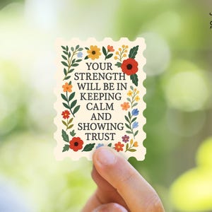 May include: A rectangular sticker with a floral border and the text "YOUR STRENGTH WILL BE IN KEEPING CALM AND SHOWING TRUST." The sticker has a cream background and is held by a hand against a blurred green background.
