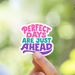 May include: A colorful sticker with the text "PERFECT DAYS ARE JUST AHEAD" in pink, teal, and purple. The sticker is held against a blurred green background. The sticker has a white border and the text is in a playful font.