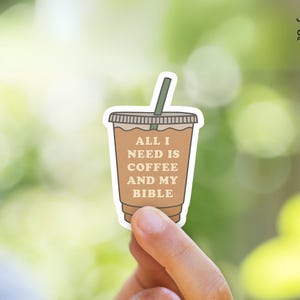 May include: A sticker featuring a coffee cup design with the text "ALL I NEED IS COFFEE AND MY BIBLE." The cup is brown with a green straw and a white outline. The background is a blurred green.