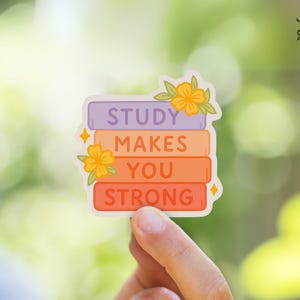 May include: A colorful sticker featuring the text "STUDY MAKES YOU STRONG" stacked on top of each other. The text is on a stack of books in shades of purple, orange, and red, with floral accents and star-shaped embellishments. The sticker is held by a hand.