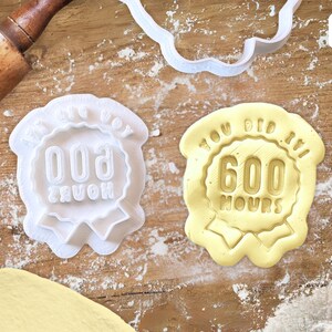 Könnte beinhalten: Zwei weiße Plastik-Ausstecher in Form eines Bandes mit dem Text "You Did It! 600 Hours" und "You Did It! 600 Hours".