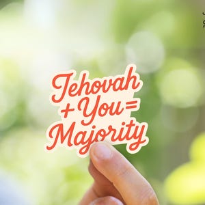 Op de afbeelding: Een sticker met de tekst "Jehovah + You = Majority" in rood scriptlettertype. De sticker heeft een crèmekleurige achtergrond en een rode rand. De achtergrond is een vage groene en witte kleur.