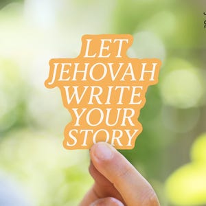 May include: An orange sticker with the text "LET JEHOVAH WRITE YOUR STORY" in white. The sticker is held by a hand against a blurred green and yellow background. The sticker has a unique shape.
