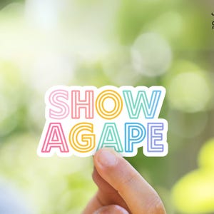 May include: A colorful sticker with the words "SHOW AGAPE" in a rainbow gradient font. The sticker has a white border and is held against a blurred green background. The sticker is from "JW Printables".