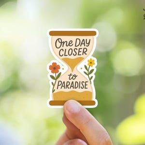 Puede incluir: Una pegatina blanca con un diseño de reloj de arena. El reloj de arena está lleno de arena amarilla y tiene el texto "One Day Closer to Paradise" escrito en él. Hay flores naranjas y amarillas a cada lado del reloj de arena.