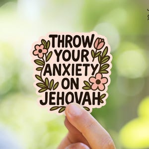 May include: A sticker with the text "THROW YOUR ANXIETY ON JEHOVAH" in black, surrounded by pink flowers and green leaves. The sticker is held by a hand against a blurred green background. The sticker has a light pink background.