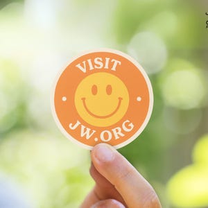 May include: A round sticker with an orange border and a yellow smiley face in the center. The words "VISIT JW.ORG" are printed in white around the smiley face. Two white dots are on either side of the smiley face.