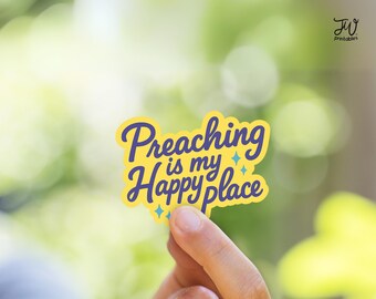 May include: A yellow sticker with the text "Preaching is my Happy place" in blue script. The sticker has a blue border and small blue star accents. The background is a blurred green.