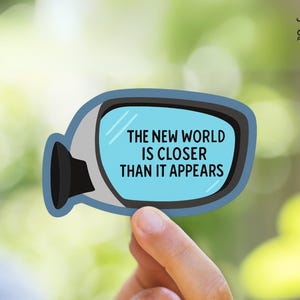 Op de afbeelding: Een blauwe en witte sticker voor de achteruitkijkspiegel van een auto met de tekst "De nieuwe wereld is dichterbij dan het lijkt".