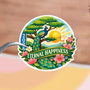 Puede incluir: Una pegatina redonda con un pavo real posado en un árbol, un sol naciente y el texto "Eternal Happiness" en una pancarta. El diseño incluye flores y los números "2026". La pegatina está sujeta por unas pinzas.
