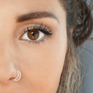 May include: Close-up portrait of a person with brown hair and brown eyes. They are wearing makeup and have a small silver nose ring.