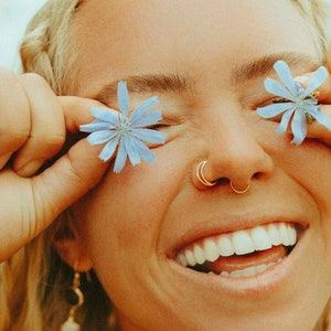 Puede incluir: Una mujer con cabello rubio sonr&iacute;e y sostiene flores azules frente a sus ojos. Lleva un aro de nariz de oro y un pendiente de oro con un colgante de perla.
