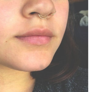 Puede incluir: Primer plano de la nariz de una persona con un anillo de septum de oro y dos aros de nariz de oro.