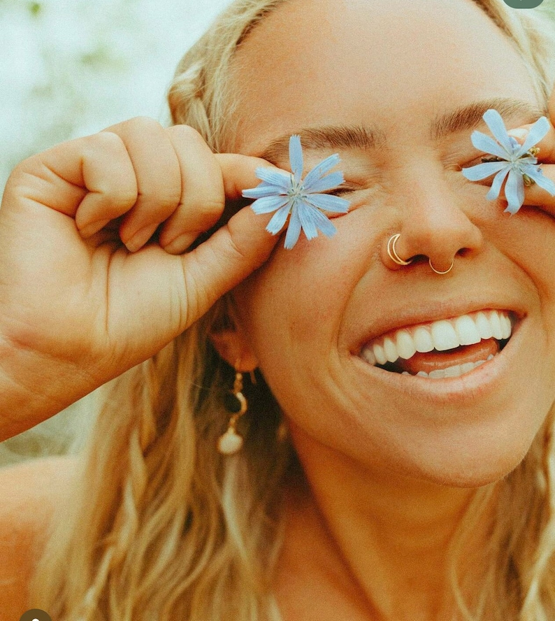 Puede incluir: Una mujer con cabello rubio sonr&iacute;e y sostiene flores azules frente a sus ojos. Lleva un aro de nariz de oro y un pendiente de oro con un colgante de perla.