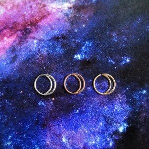 Puede incluir: Tres anillos de nariz en forma de media luna en plata, oro rosa y oro. Los anillos se muestran sobre un fondo con estampado de galaxia.