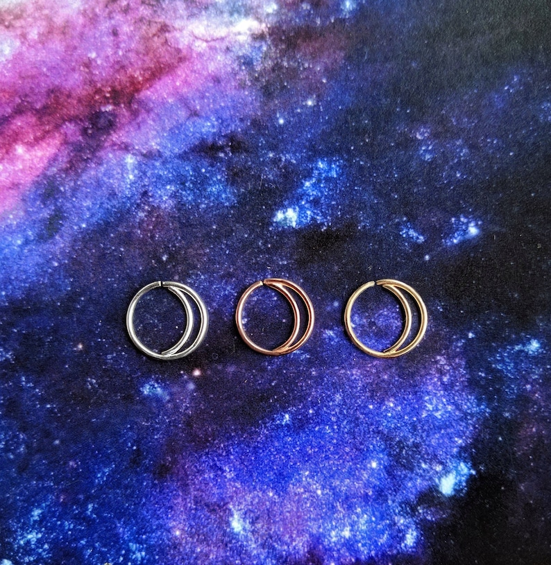 Puede incluir: Tres anillos de nariz en forma de media luna en plata, oro rosa y oro. Los anillos se muestran sobre un fondo con estampado de galaxia.