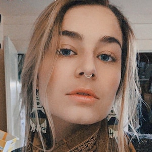 Puede incluir: Un retrato de cerca de una persona con cabello rubio y ojos azules. Lleva una bufanda de color marr&oacute;n con estampado y tiene un aro de nariz plateado.