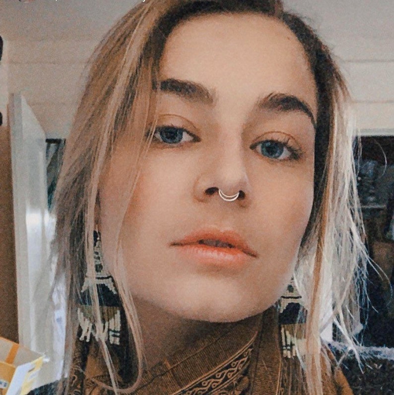 Puede incluir: Un retrato de cerca de una persona con cabello rubio y ojos azules. Lleva una bufanda de color marr&oacute;n con estampado y tiene un aro de nariz plateado.