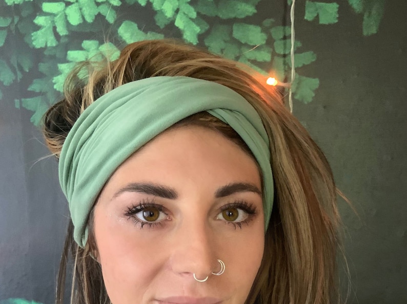Puede incluir: Una diadema el&aacute;stica de color verde salvia puesta en la cabeza de una persona. La diadema est&aacute; retorcida y anudada en la parte superior.