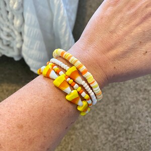May include: A set of four bracelets featuring a candy corn design. The bracelets are made of white, orange, and yellow beads. The candy corn design is made of white and yellow beads.