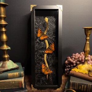 May include: A vertical black rectangular shadow box frame with gold corner accents. Inside, a gold-colored snake sculpture winds upward, adorned with three orange and black butterflies against a black embossed floral background.