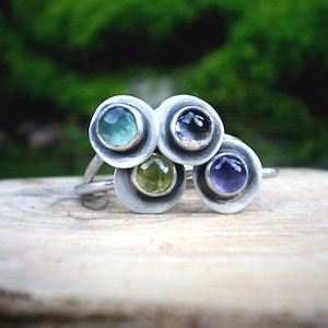 May include: A silver ring with four round gemstones set in a cluster. The gemstones are green, blue, purple, and clear.