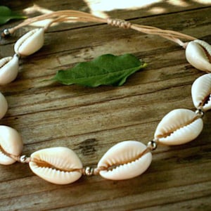 May include: A necklace composed of white cowrie shells, strung on a beige cord, with small silver beads connecting the shells. The necklace is presented on a wooden surface, accompanied by a green leaf.