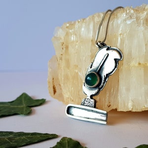 Puede incluir: Un collar de plata con un colgante con forma de pluma y tintero. La pluma tiene una piedra preciosa verde en el centro.