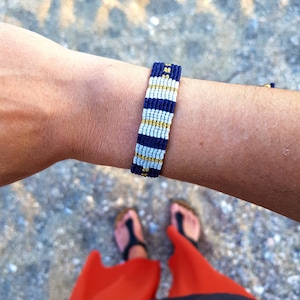 May include: A blue and white woven bracelet with gold accents. The bracelet is worn on a person's wrist.
