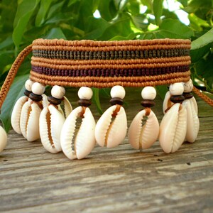 Puede incluir: Un brazalete de cordón trenzado marrón, verde y morado con conchas de cauri blancas colgando de él. El brazalete está sobre una superficie de madera.