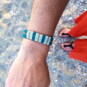 May include: A turquoise and white friendship bracelet with gold accents. The bracelet is woven with a simple pattern and is tied around a person's wrist.