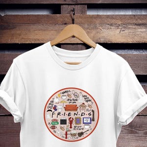 Könnte beinhalten: Weißes T-Shirt mit einem kreisförmigen Grafik mit verschiedenen Symbolen und Texten, die sich auf die Fernsehserie "Friends" beziehen. Die Grafik enthält die Wörter "Friends" und "How you doin'" sowie Bilder einer violetten Tür, einer Kaffeetasse, eines Hummers, eines Fußballs und eines Sofas.
