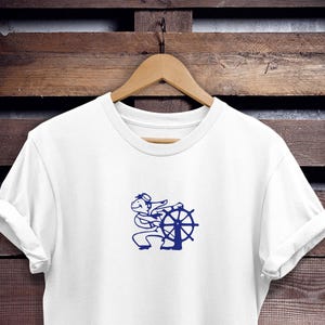May include: White t-shirt with a navy blue graphic of a cartoon sailor steering a ship's wheel. The shirt is hanging on a wooden hanger against a dark wood background. The design features a sailor with a hat and a ship's wheel.