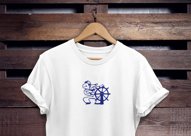 May include: White t-shirt with a navy blue graphic of a cartoon sailor steering a ship's wheel. The shirt is hanging on a wooden hanger against a dark wood background. The design features a sailor with a hat and a ship's wheel.