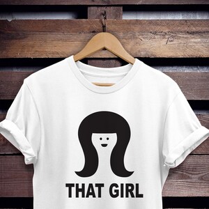 May include: White t-shirt with a black silhouette of a woman with a bob haircut and the text "THAT GIRL" printed on the front.