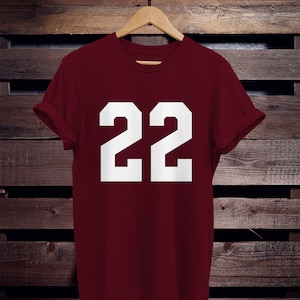 K&ouml;nnte beinhalten: Ein weinrotes T-Shirt mit der wei&szlig;en Zahl "22" in fetter Blockschrift. Das Shirt h&auml;ngt an einem Holzb&uuml;gel vor einem dunklen Holzgrund. Die &Auml;rmel sind hochgekrempelt.