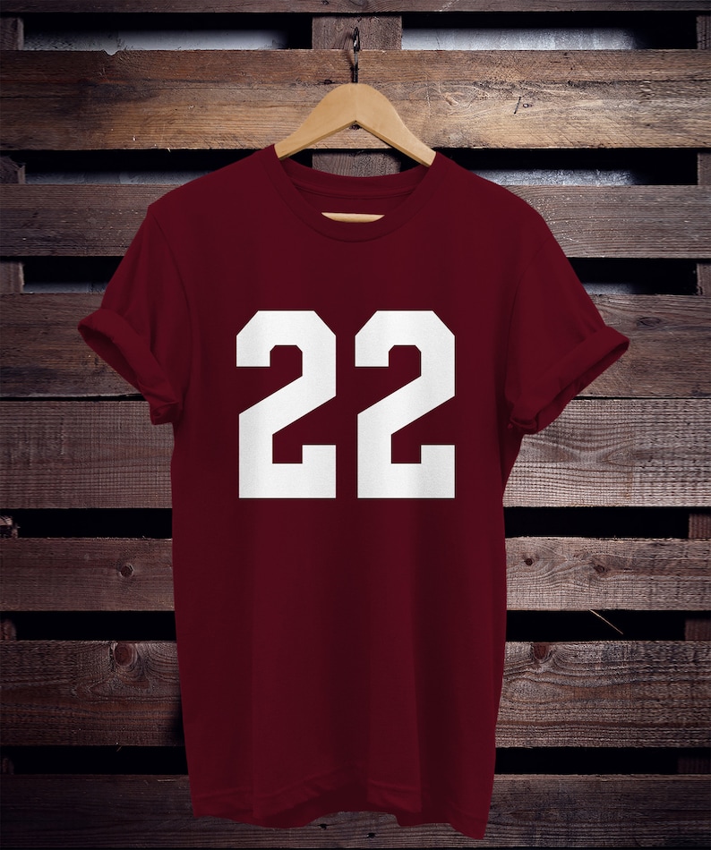 K&ouml;nnte beinhalten: Ein weinrotes T-Shirt mit der wei&szlig;en Zahl "22" in fetter Blockschrift. Das Shirt h&auml;ngt an einem Holzb&uuml;gel vor einem dunklen Holzgrund. Die &Auml;rmel sind hochgekrempelt.