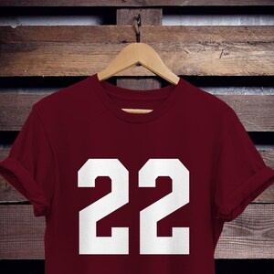 K&ouml;nnte beinhalten: Ein weinrotes T-Shirt mit der wei&szlig;en Zahl "22". Das T-Shirt h&auml;ngt an einem Holzb&uuml;gel vor einem Hintergrund aus Holzbrettern. Die &Auml;rmel sind hochgekrempelt.