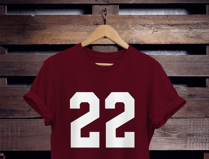 K&ouml;nnte beinhalten: Ein weinrotes T-Shirt mit der wei&szlig;en Zahl "22". Das T-Shirt h&auml;ngt an einem Holzb&uuml;gel vor einem Hintergrund aus Holzbrettern. Die &Auml;rmel sind hochgekrempelt.