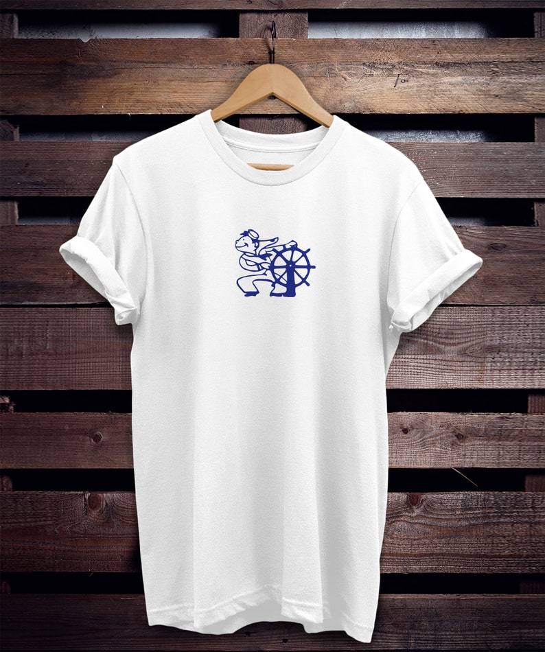 May include: White t-shirt with a blue graphic of a sailor at a ship's wheel. The shirt is hanging on a wooden hanger against a dark brown wooden background. The sleeves are rolled up.