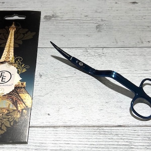 Puede incluir: Un par de cortamechas de vela de metal azul con un mango curvo. Los cortamechas están sobre una superficie de madera clara. El embalaje de los cortamechas está en el fondo y presenta una imagen de la Torre Eiffel.