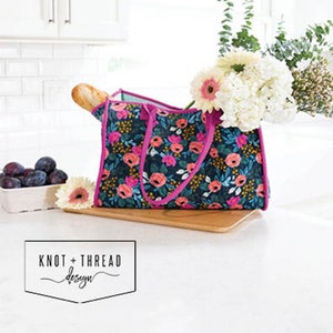 May include: A tote bag with a floral pattern in pink, blue, and yellow, with the text "ALL THE THINGS TOTE". The bag is filled with flowers and a loaf of bread. The bag has a pink trim and handles. The logo "KNOT + THREAD design" is in the bottom left corner.