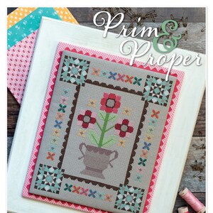Op de afbeelding: Een kruissteekpatroon met de titel "Prim & Proper" met een bloemenontwerp met een vaas en een bruine, witte en blauwe rand. Het patroon is ingelijst en wordt weergegeven op een houten oppervlak met spoelen garen en een schaar.