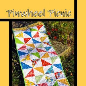 May include: A colorful quilt with a pinwheel design. The quilt is made with a variety of fabrics, including floral prints, gingham, and solid colors. The quilt is laid out on a rock in a grassy area. The quilt is titled "Pinwheel Picnic" and is by Tricia Lynn Maloney, Orphan Quilt Designs for Villa Rosa Designs.