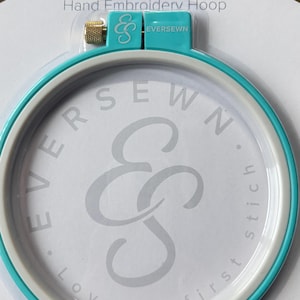 May include: A 7-inch EverSewn hand embroidery hoop. The hoop has a teal outer ring and a white inner ring. The center features the EverSewn logo and text. The text "Love at first stitch" is also visible.