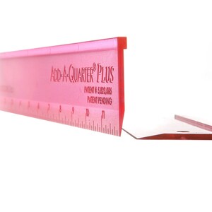 May include: Pink plastic ruler with the text "Add-A-Quarter® Plus" and "Patent # 5,823,086 Patent Pending". The ruler is 12 inches long and has markings for inches and fractions of an inch.